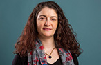A woman (Adelina Feo) with long, curly brown hair and hazel eyes, weraing a flower pattern scarf and gold necklace, looking at the camera with a slight grin.